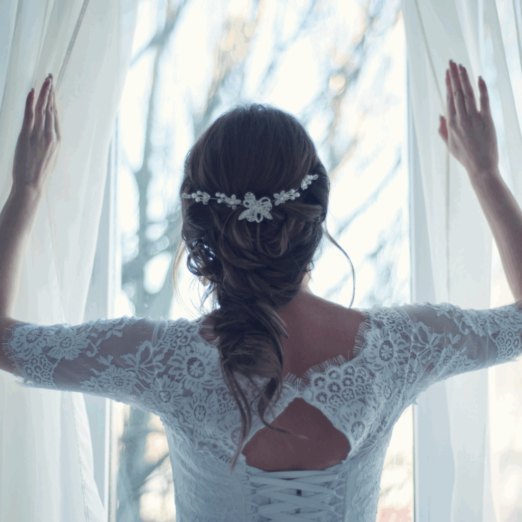 Bride in lace gown opening white curtains, showcasing an elegant floral hairpiece and relaxed wedding-morning vibe