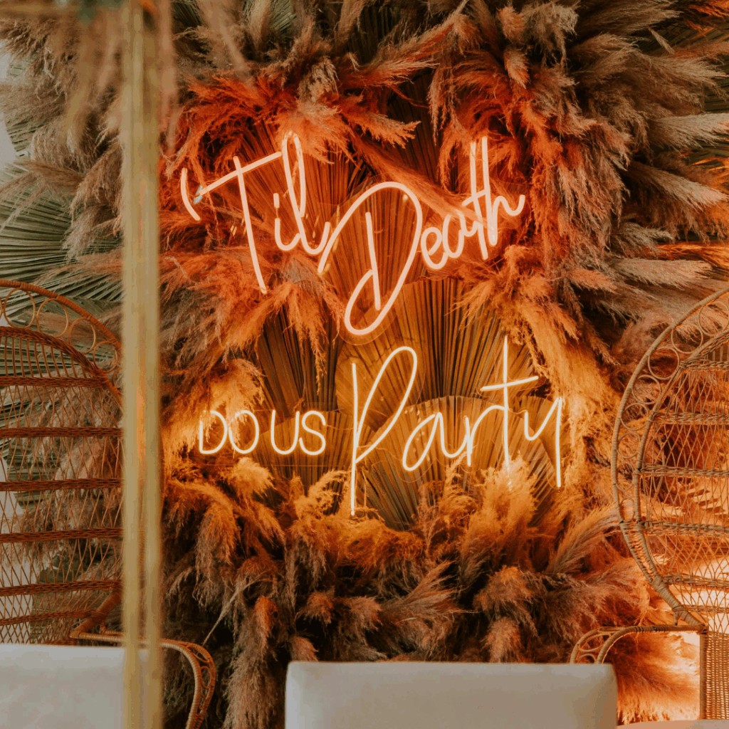 Boho wedding backdrop with pampas grass, dried palm leaves, and neon sign reading “’Til Death Do Us Party.