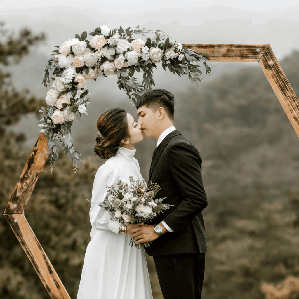 Bride and groom sharing a kiss under a rustic hexagon arch adorned with blush and ivory flowers during an outdoor wedding ceremony.
