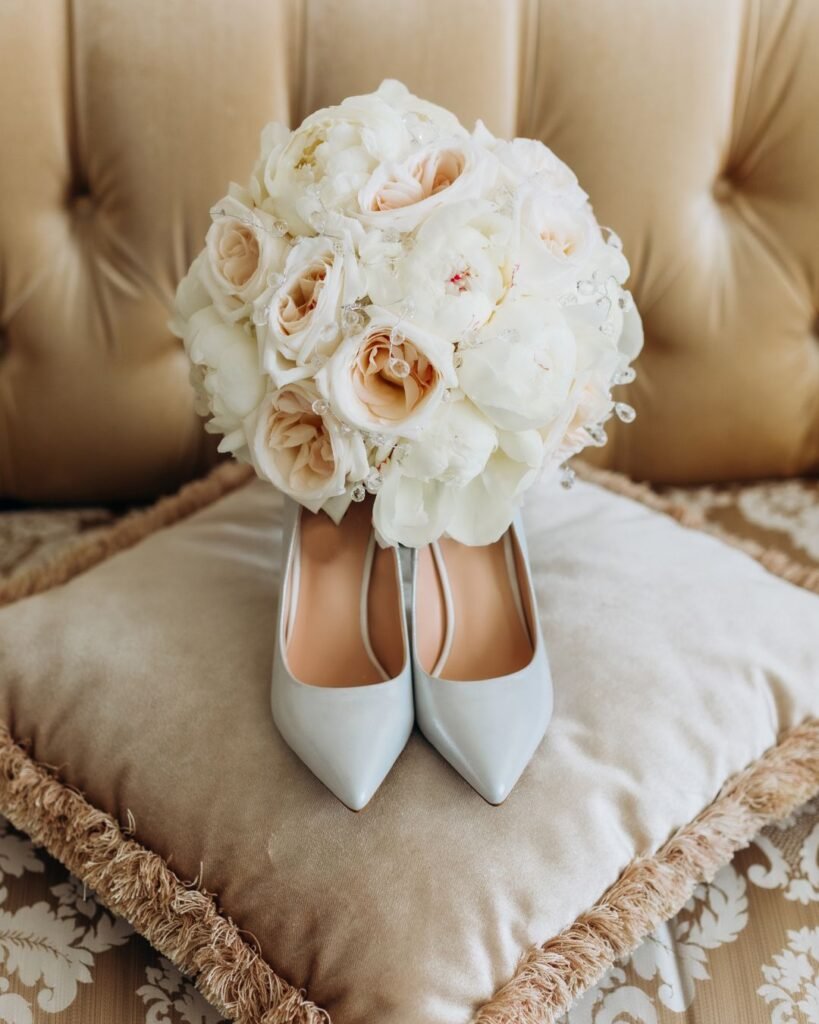  Elegant bridal bouquet with ivory and blush roses placed on top of white wedding heels, resting on a plush velvet pillow.