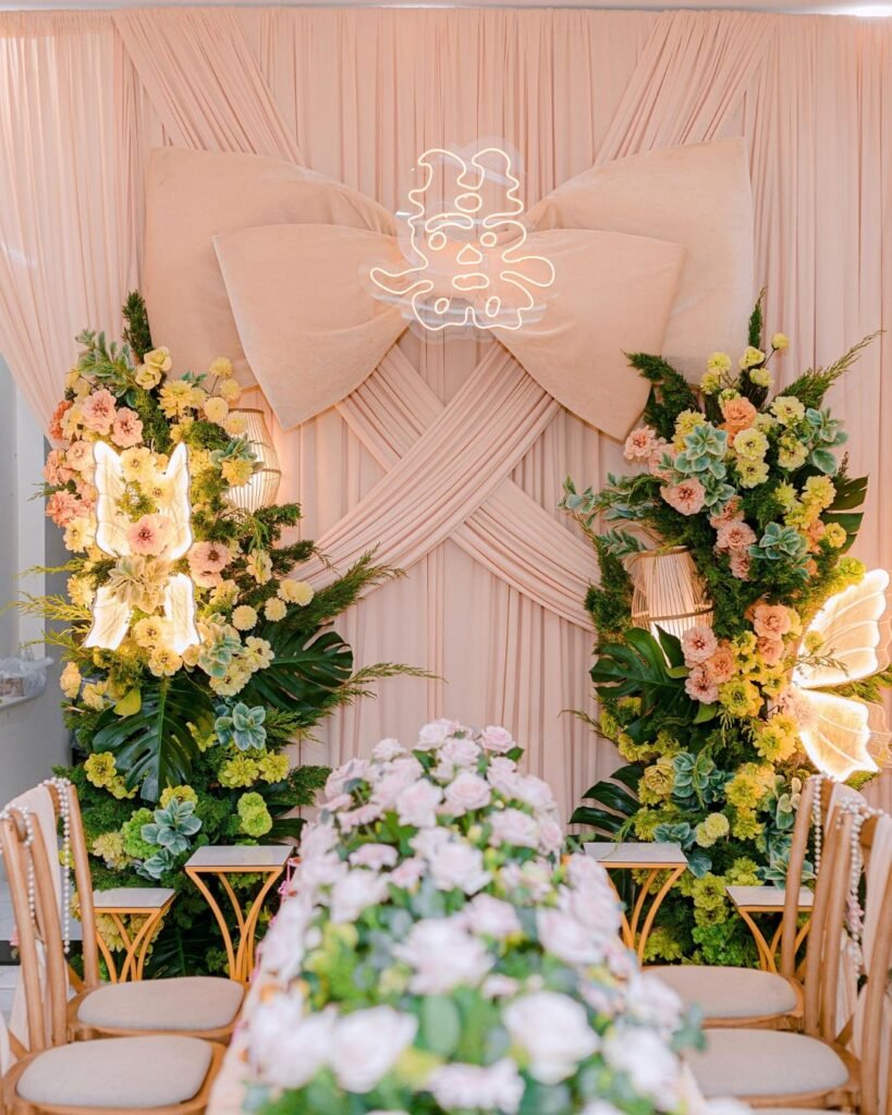 “Elegant wedding reception table setup with lush floral arrangements, pastel roses, and a giant fabric bow backdrop with neon light art.”

