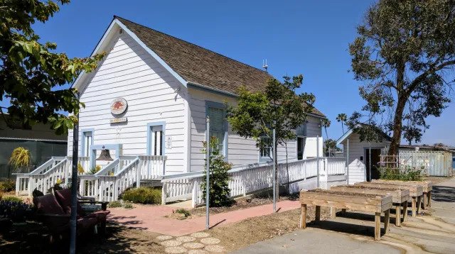Side view of the historic 1883 Schoolhouse in Encinitas, California, with ramp and shade trees beneath a clear blue sky