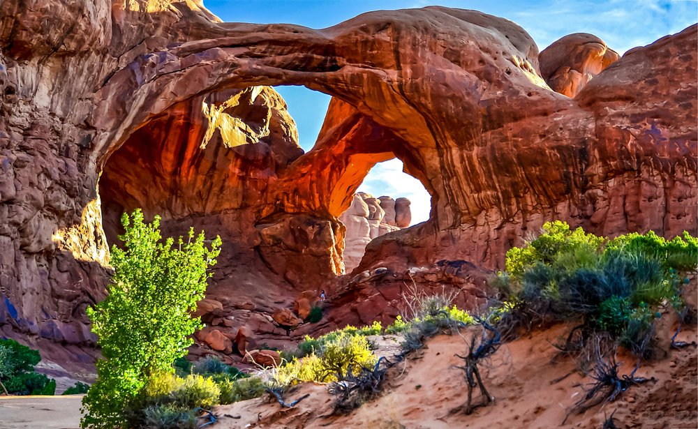 Sunlight pouring through a towering sandstone arch with desert shrubs in the foreground