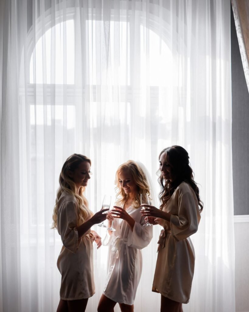 Three women in personalized bridal robes toasting with champagne in front of a sheer window