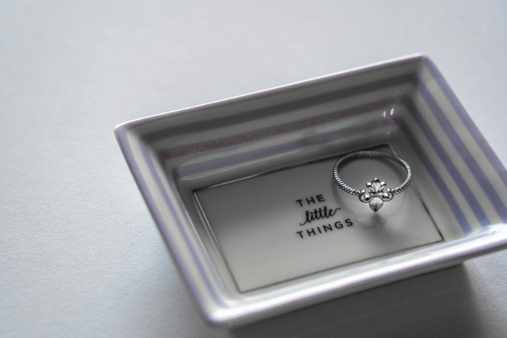 Silver engagement ring displayed on a striped wedding ring dish with the words “The little things.”