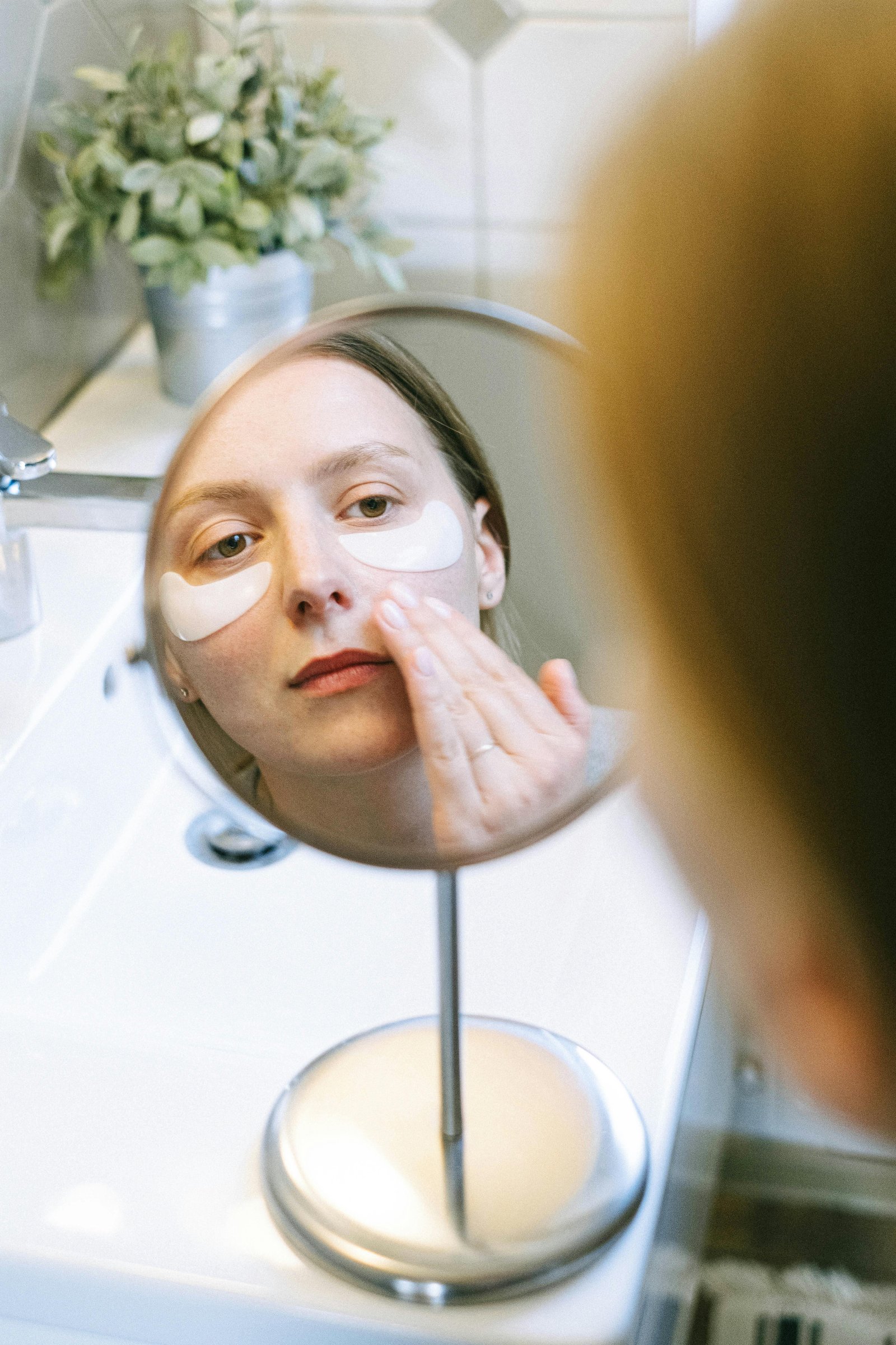Woman applying under-eye patches while looking in a mirror as part of her skincare routine.