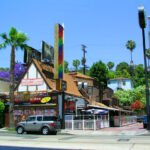 Street view of Rainbow Bar & Grill on Sunset Strip in West Hollywood, framed by palm trees and its rainbow sign.