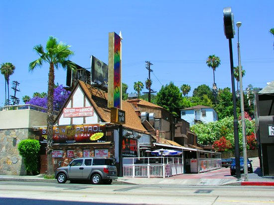 Street view of Rainbow Bar & Grill on Sunset Strip in West Hollywood, framed by palm trees and its rainbow sign.