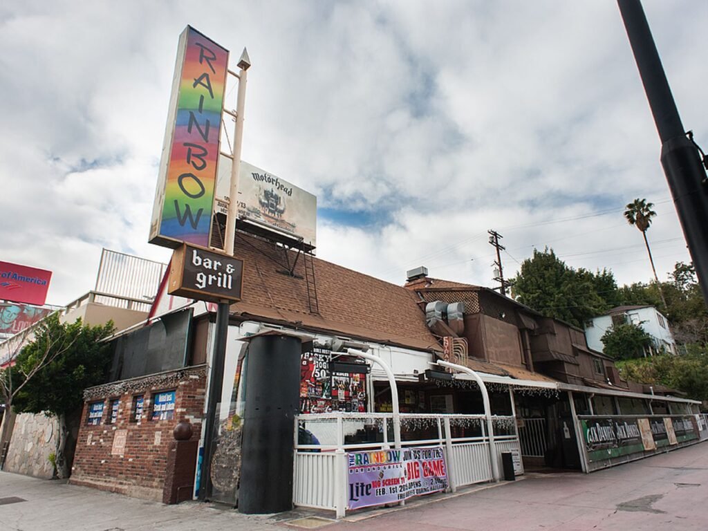 Close-up of the iconic Rainbow Bar & Grill sign and brick facade in West Hollywood beneath a cloudy sky.