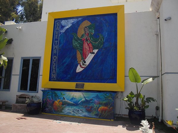Wide view of the Surfing Madonna mosaic in a yellow frame on a white wall in Encinitas, with an ocean-themed mural beneath.