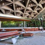 Sunken living room of the Sheats-Goldstein House featuring a triangular concrete ceiling, built-in brown leather banquette, and floor-to-ceiling glass overlooking lush foliage