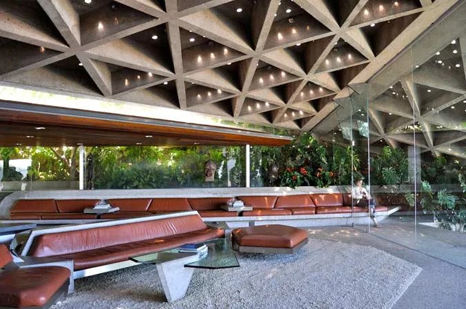 Sunken living room of the Sheats-Goldstein House featuring a triangular concrete ceiling, built-in brown leather banquette, and floor-to-ceiling glass overlooking lush foliage