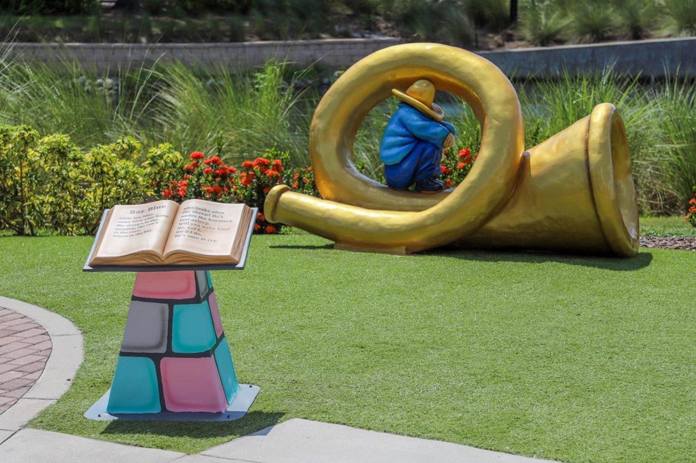 Boy Blue statue from Ulele Fairyland Figures Tampa crouched inside a giant golden horn beside an open storybook pedestal on the Riverwalk