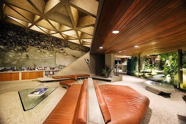 Evening interior view of the Sheats-Goldstein House with illuminated coffered ceiling, angled wood paneling, and curved brown leather seating around a glass coffee table