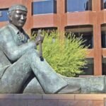 Bronze sculpture of a seated Navajo Code Talker holding a traditional message stick at Phoenix Plaza, with a red office building and desert greenery behind
