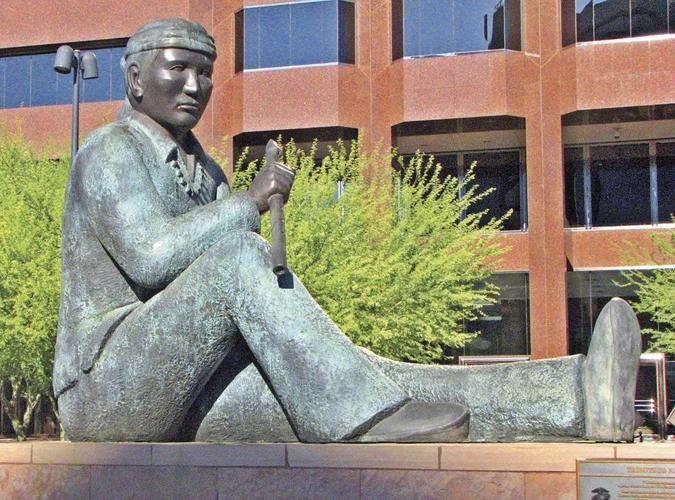 Bronze sculpture of a seated Navajo Code Talker holding a traditional message stick at Phoenix Plaza, with a red office building and desert greenery behind