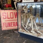 A skeleton display next to a vintage red “SLOW FUNERAL” sign at Dysfunctional Grace Tampa, featuring antique medical and anatomical decor.