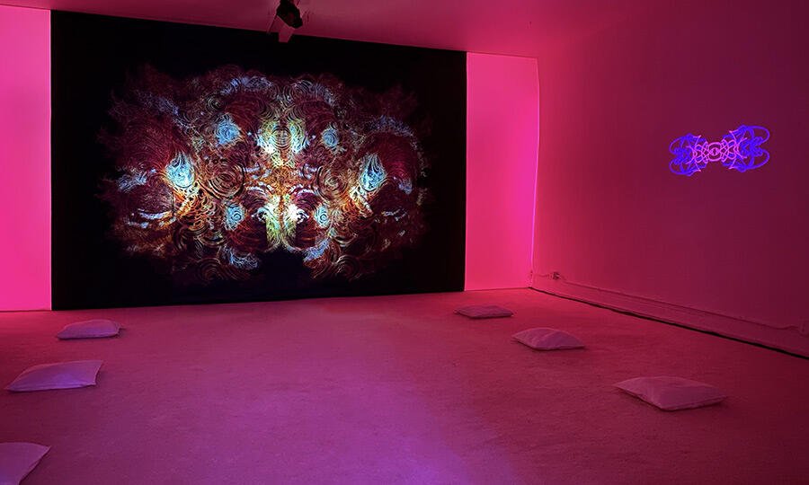 Dream House New York City room glowing pink, kaleidoscopic projection on black wall, white cushions on floor.