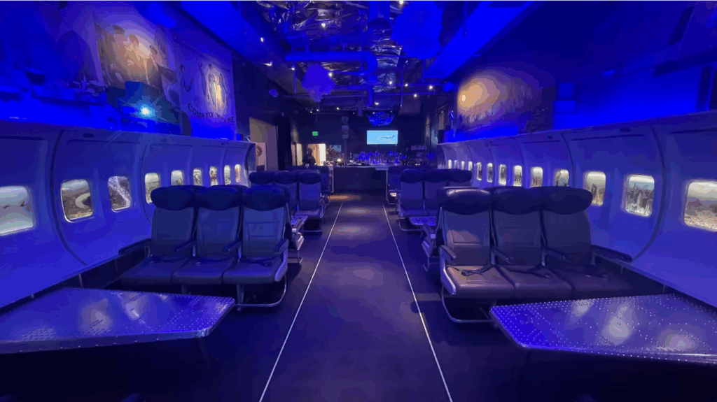 Central aisle view of PS Air Bar Palm Springs, featuring airline cabin seats, window fuselage panels and blue mood lighting.