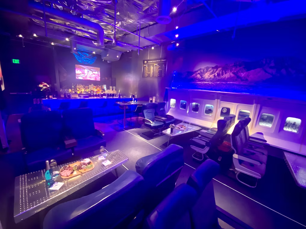 Angled view inside PS Air Bar Palm Springs with airplane seats, back-lit window walls, charcuterie on a wing table and bar.