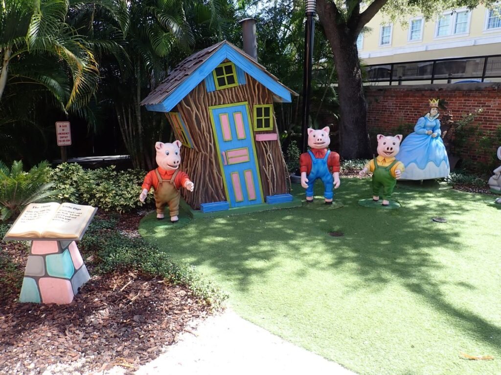 Three Little Pigs display at Ulele Fairyland Figures Tampa with a crooked wooden house, three pig statues in colorful outfits, and an open storybook podium