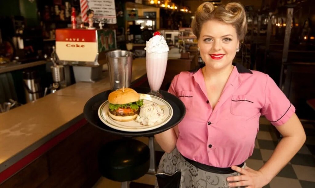 Retro-styled server holding a burger, potato salad, and strawberry milkshake inside MacAlpine’s Soda Fountain Phoenix.