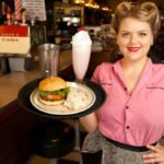 Retro-styled server holding a burger, potato salad, and strawberry milkshake inside MacAlpine’s Soda Fountain Phoenix.