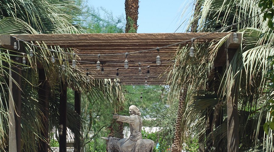 Statue of Jesus along “The Walk” sculpture garden at Shields Date Garden Indio, framed by palm fronds and a wooden arbor