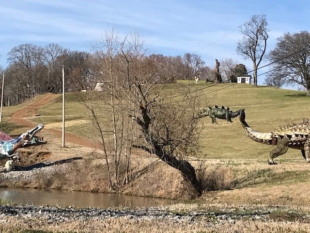 Ankylosaurus and other dinosaur statues on a grassy hillside beside a creek at Boedecker Sculpture Garden in Nashville.