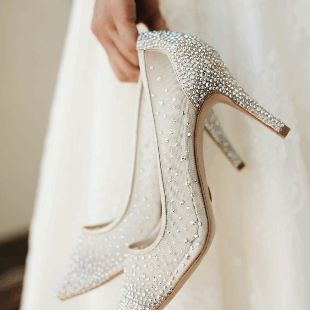 Bride holding rhinestone-covered stiletto heels, sparkling bridal shoes against lace gown