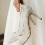 Bride holding rhinestone-covered stiletto heels, sparkling bridal shoes against lace gown