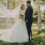 Bride in an off-shoulder lace gown and groom in a navy tux posing outdoors with veil billowing—a nod to Barcelona bridal trends’ romantic elegance