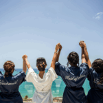 Bridal party in matching silk robes (Bride, Maid of Honor, Bridesmaid) raising hands overlooking a seaside view