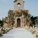 Wedding ceremony aisle decorated with pampas grass and dried greenery leading to a stone chapel with boho floral arch.