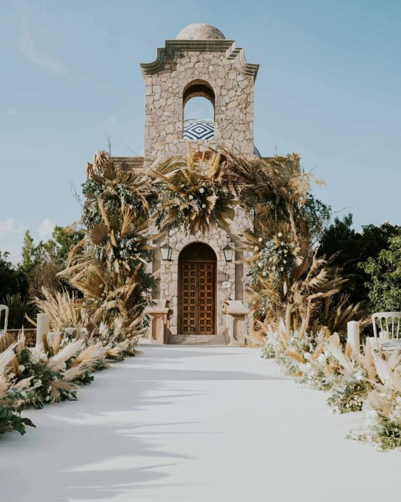 Wedding ceremony aisle decorated with pampas grass and dried greenery leading to a stone chapel with boho floral arch.