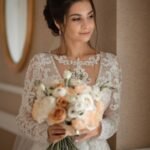 Bride holding soft peach and ivory bouquet, wearing a lace wedding gown with elegant bridal makeup and loose updo hairstyle