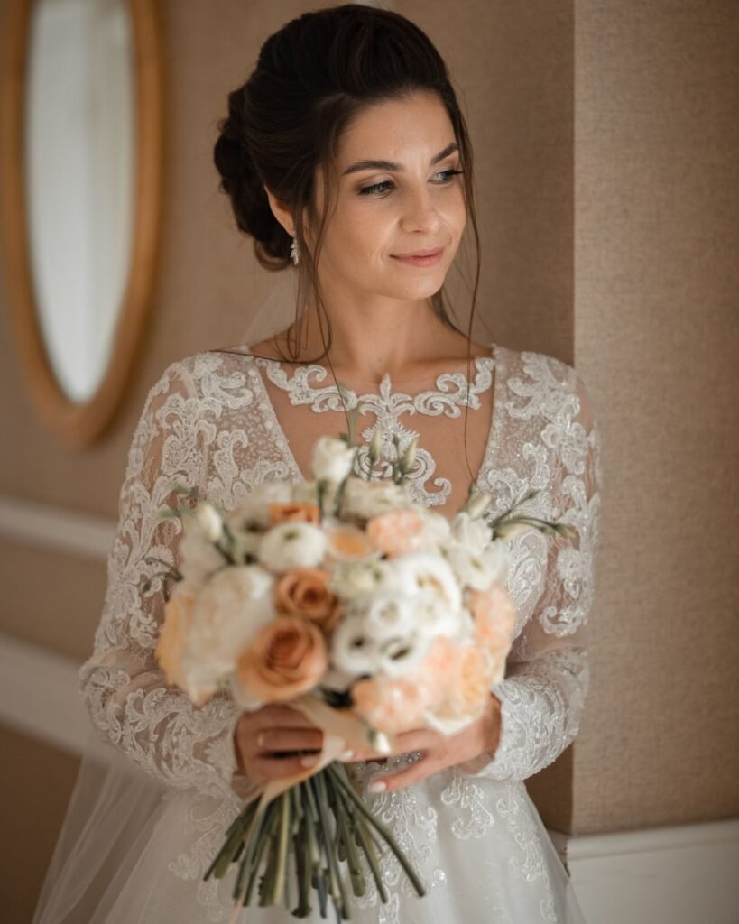 Bride holding soft peach and ivory bouquet, wearing a lace wedding gown with elegant bridal makeup and loose updo hairstyle