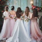 Bride and bridesmaids in pastel dresses holding floral bouquets, standing with their backs to the camera in an elegant vintage venue.