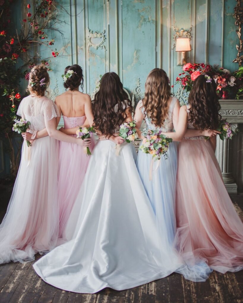 Bride and bridesmaids in pastel dresses holding floral bouquets, standing with their backs to the camera in an elegant vintage venue.