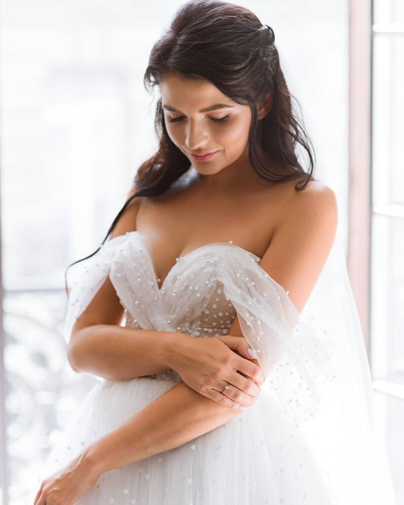 “Bride in a tulle wedding gown adjusting her arm for picture-perfect wedding prep in natural light.”