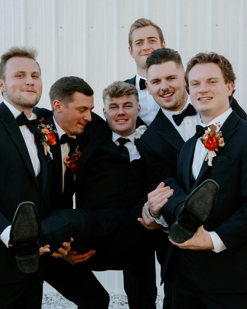 Creative groomsmen gift swap moment featuring six groomsmen in tuxedos lifting the groom for a playful photo