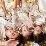 Bridesmaids in satin robes laughing on bed with confetti during bridal party getting ready playlist moment