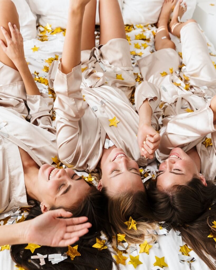 Bridesmaids in satin robes laughing on bed with confetti during bridal party getting ready playlist moment