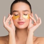 Woman with eyes closed applying gold under eye patches for skincare and puffiness reduction