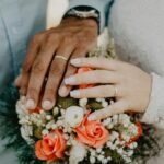 “Close-up of newlywed couple’s hands with wedding rings resting on a bouquet of coral and white roses.”