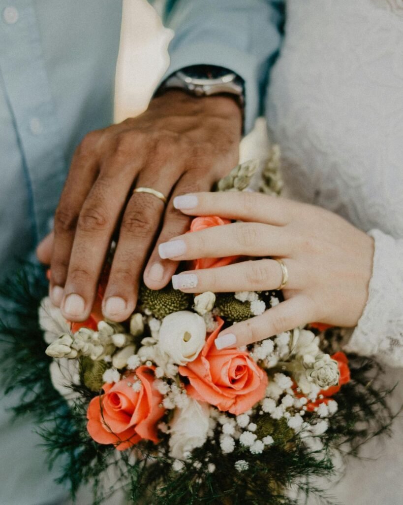 “Close-up of newlywed couple’s hands with wedding rings resting on a bouquet of coral and white roses.”
