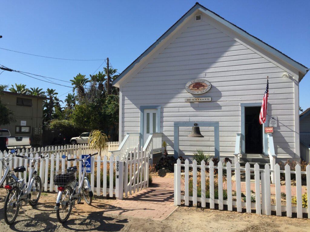 Front entrance of the 1883 Schoolhouse in Encinitas, CA, featuring its bell, white picket fence, and American flag on a sunny day