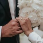 Close-up of bride and groom holding hands with wedding rings and white bridal bouquet in the background.