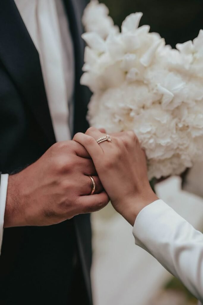 Close-up of bride and groom holding hands with wedding rings and white bridal bouquet in the background.