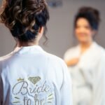 Back view of a personalized bridal robe in white satin embroidered with ‘Bride to Be’ and gold accents